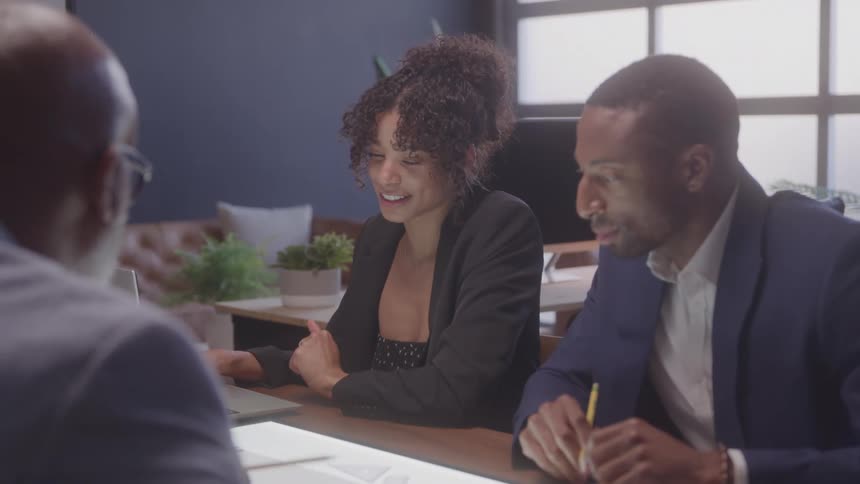 Three professionals two men and a woman seated at a conference table with the woman in the center laughing and gesturing as they engage in a collaborative discussion