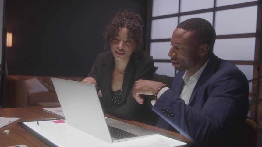 A woman and a man in business attire sit side by side at a laptop leaning in closely as they discuss something on the screen