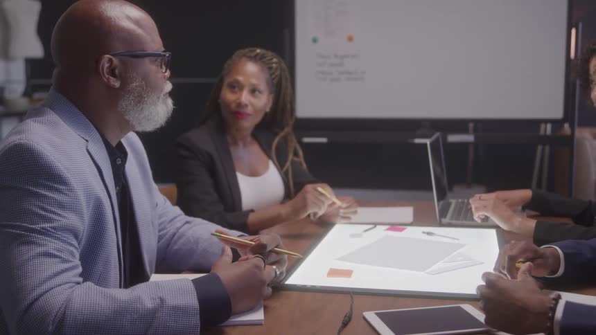 A man and a woman in business attire sit across from each other at a table with documents and laptops engaged in a professional discussion