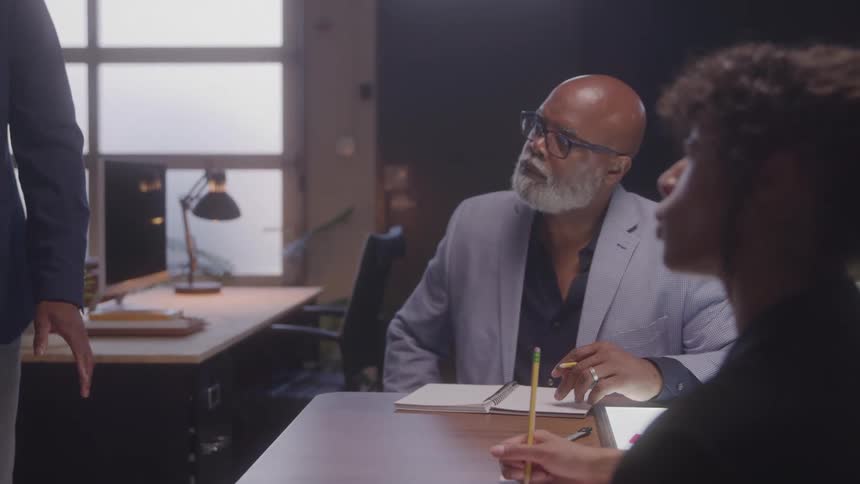 A professional man in glasses listening intently to a person offscreen while seated at a desk with an open notebook in a dimly lit office