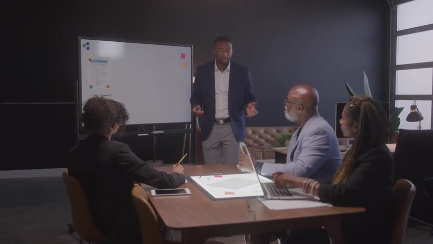 Man in blazer standing in an office room talking to other people who are sitting at a table