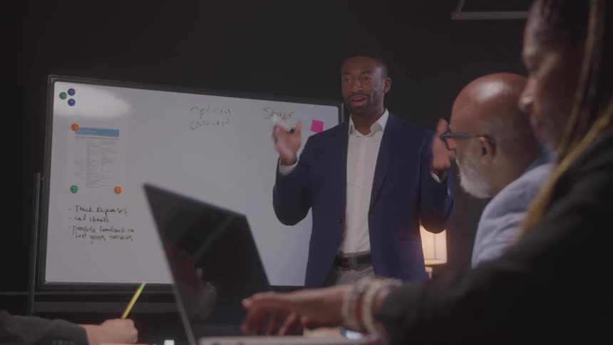 A black man in a blue suit stands in front of a whiteboard gesturing with his hands as he presents to a group of diverse colleagues seated around a table with laptops and papers in front of them