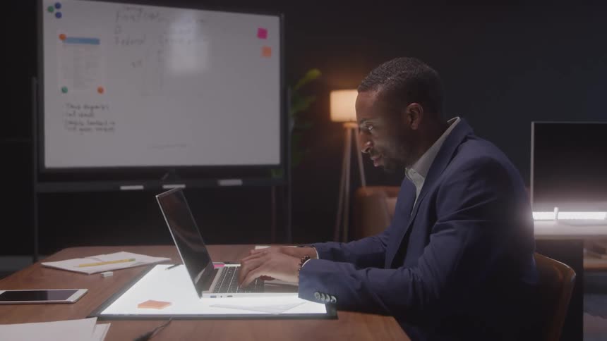 A young man sits at a desk typing on a computer wearing a blue blazer suit with a projector screen nearby inside an office