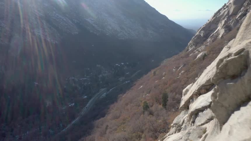 A climber ascends a steep sunlit cliff face with a deep valley and winding road visible far below