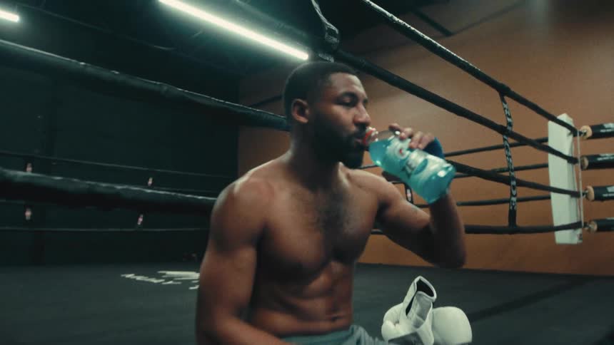 A shirtless man sits in a boxing ring drinking from a blue sports drink bottle during a break in training