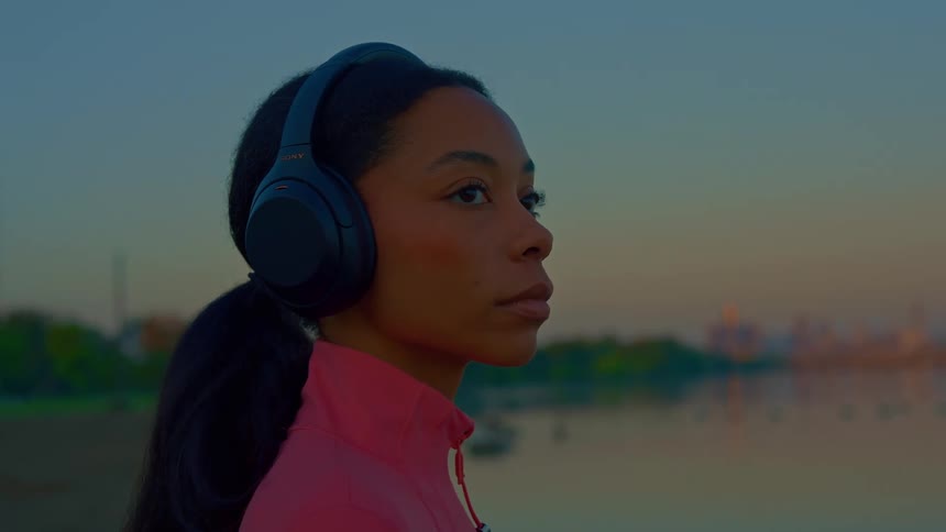 Woman wearing headphones looks at the horizon by the water at sunset
