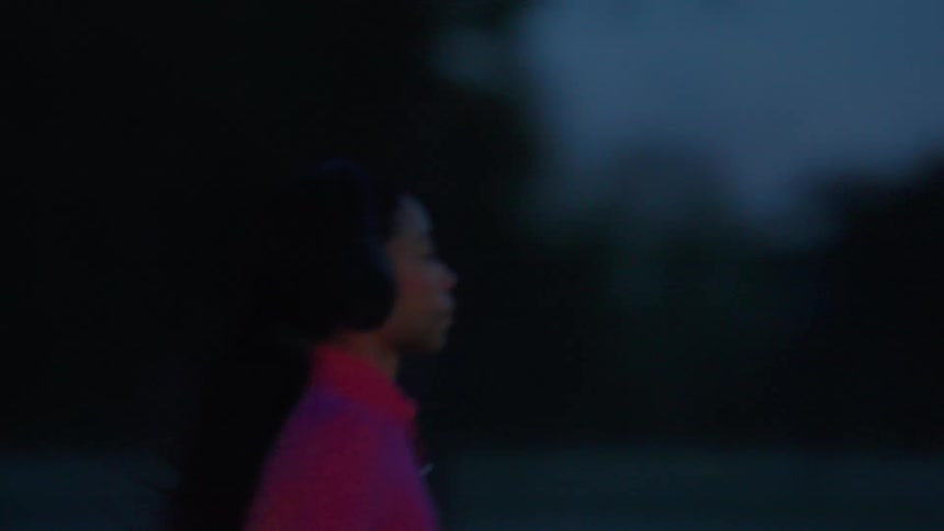 A young woman wearing headphones jogs across a field outdoors at dusk