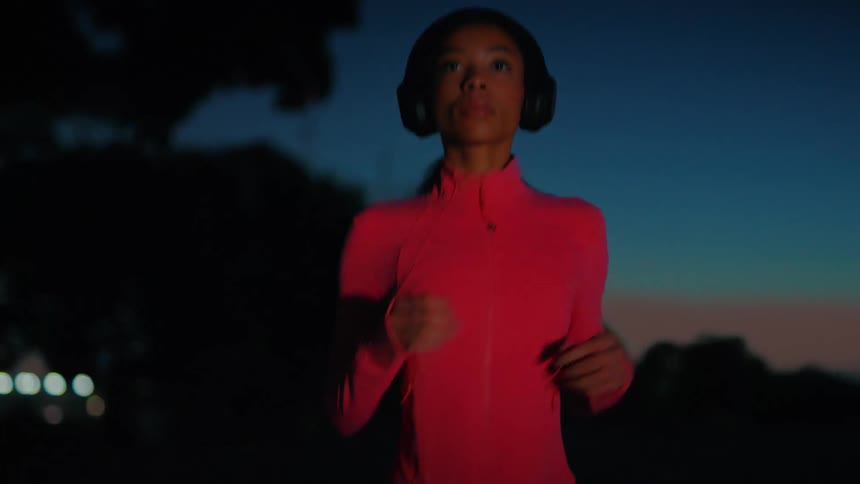A young black woman wearing headphones jogs on a street outdoors in the evening