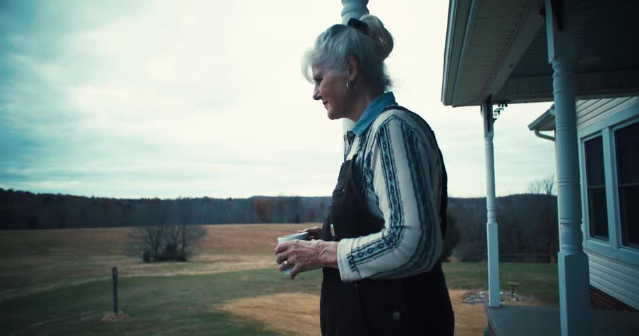 Elderly woman stands on porch of remote country house with a cup of coffee in the morning