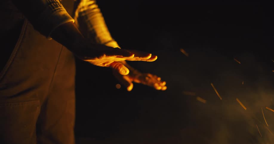 A man stands by a fire at night holding out his hands to warm them