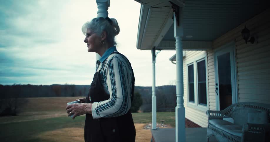Elderly woman gazes at fields from a porch holding a coffee cup