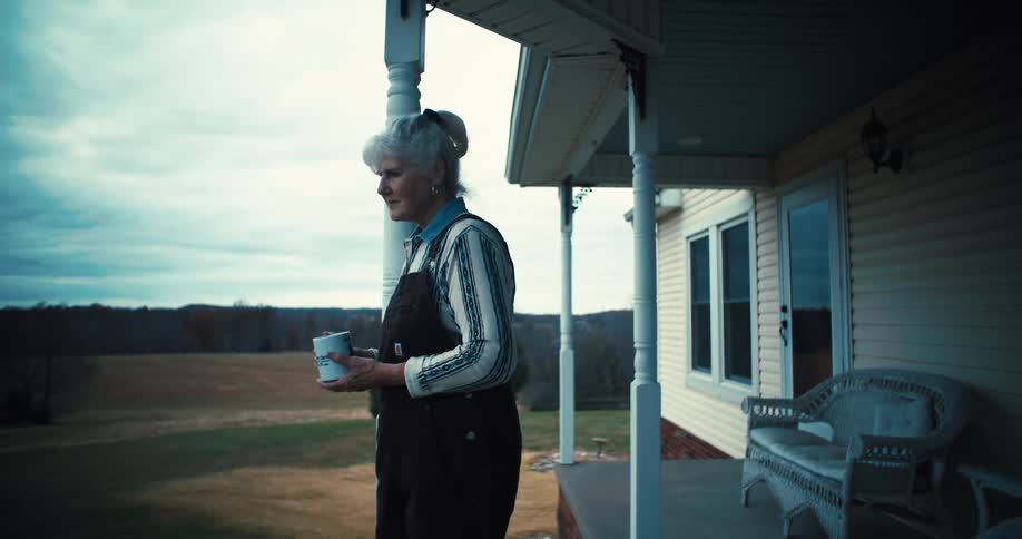 A sad looking elderly woman holding a mug as she stands in front of her porch