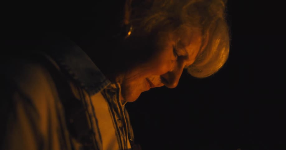 An older woman with a somber expression her face dramatically lit from one side against a completely dark background