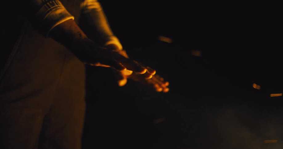 A persons hands are extended toward a warm light source likely a fire casting an orange glow against the dark surroundings