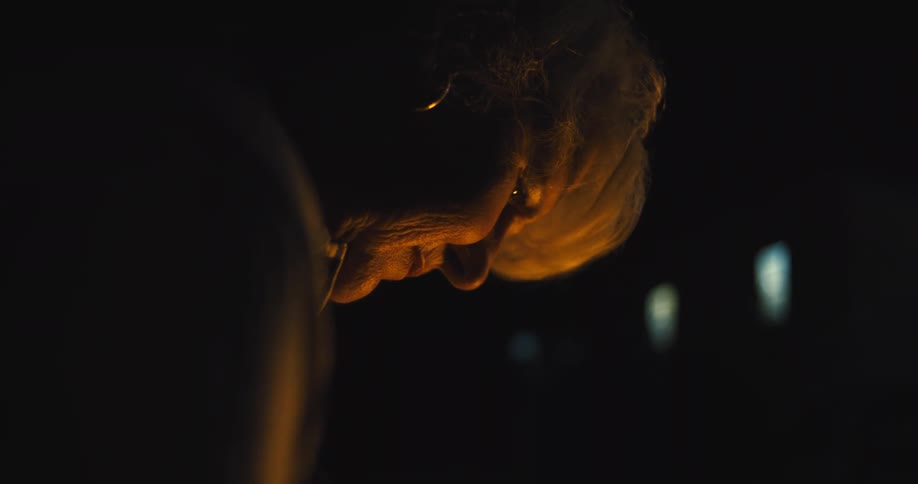 A close view shows the face of a woman with white hair and a hoop earring with her head tilted down as a yellow light illuminates her from the side