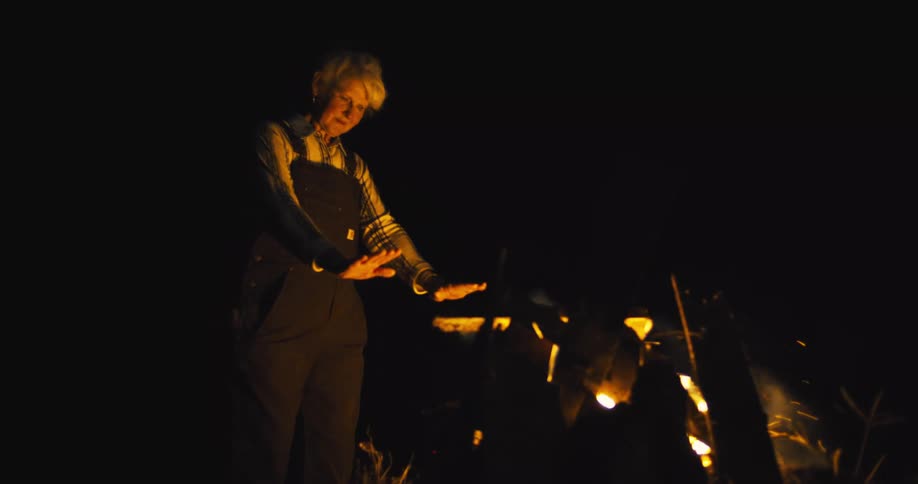 Elderly woman standing over a bonfire holding out her hands for warmth