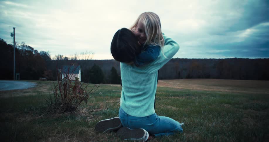 A mother hugs her daughter warmly in the grass as a dog walks past them in the background