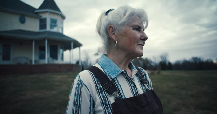 Elderly woman stands in the large yard surrounding her large rural home and looks around on an overcast day