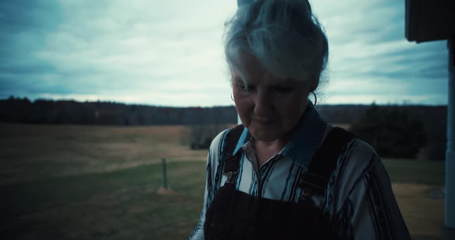 The video shows a medium shot of an elderly woman with grey hair tied in a ponytail wearing a striped shirt and overalls she is standing on a porch looking down with a rural landscape in the background under a cloudy sky