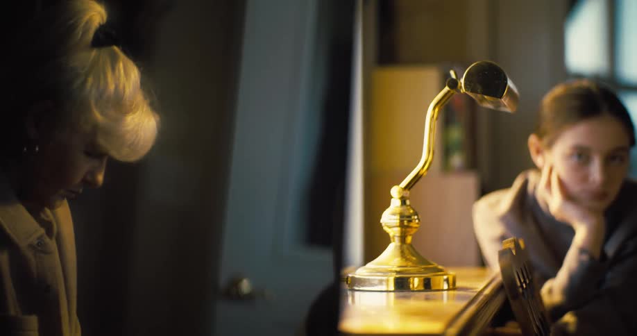 The video shows two people sitting at a table with a goldencolored desk lamp between them on the left an older person with their hair in a whiteblonde bun is seen in profile on the right a young woman rests her chin on her hand looking thoughtfully