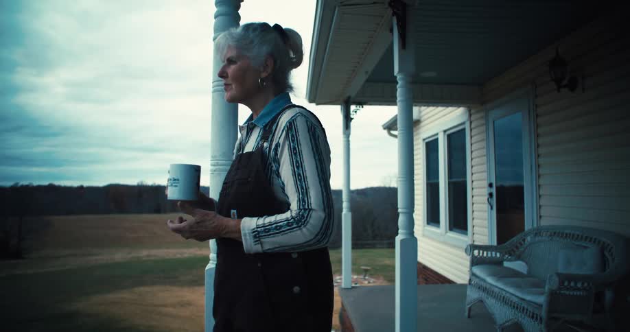 Elderly woman standing on porch at her remote country house drinking coffee at sunrise