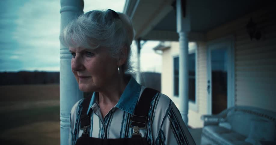 An older woman with a pensive expression wearing a striped shirt and overalls standing on a rustic porch and looking off into the distance
