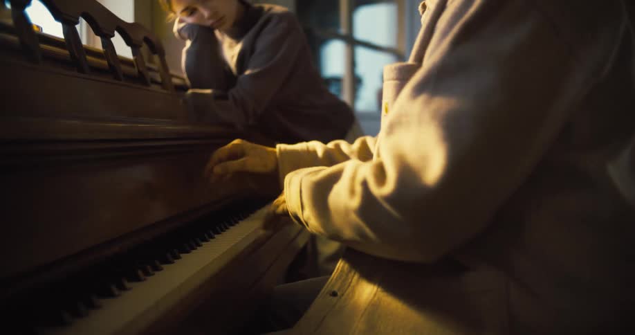 A student rests her chin on her hand looking intently at an instructor who is playing a wooden piano in a dimly lit room