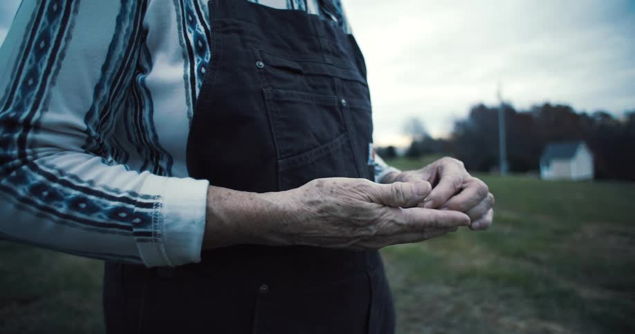 A farmer wearing dark overalls over a striped long sleeve shirt holds a ring in their wrinkled hands looking down at it then puts it into a pocket
