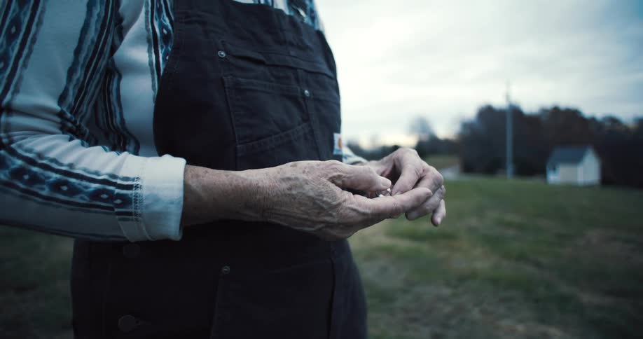 A farmer wearing dark overalls over a striped long sleeve shirt holds a golden ring in their wrinkled hands then puts it into a pocket