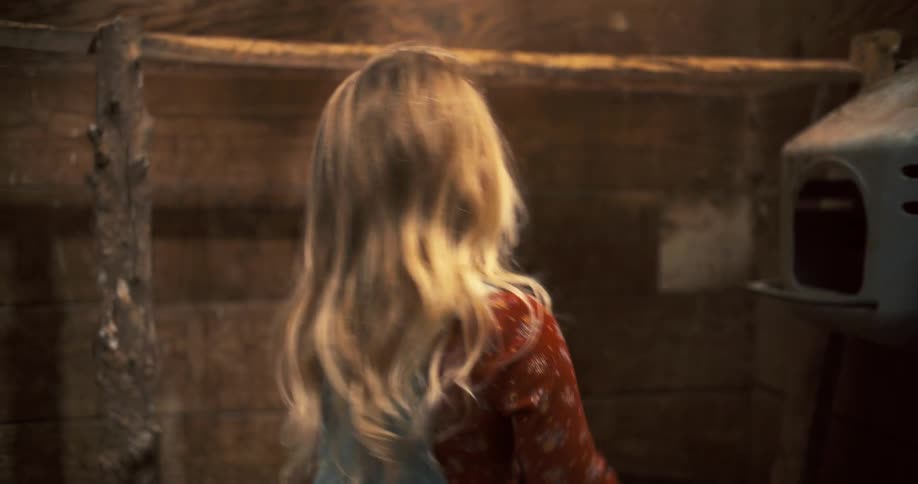 A girl with long light colored hair and wearing denim overalls over a red shirt looks at a row of chicken coops in a dark barn points her index finger toward the coops then a chicken sticks its head out of the one she is pointing at