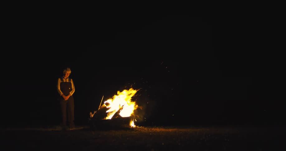 A woman stands beside a burning log of wood outdoors at night