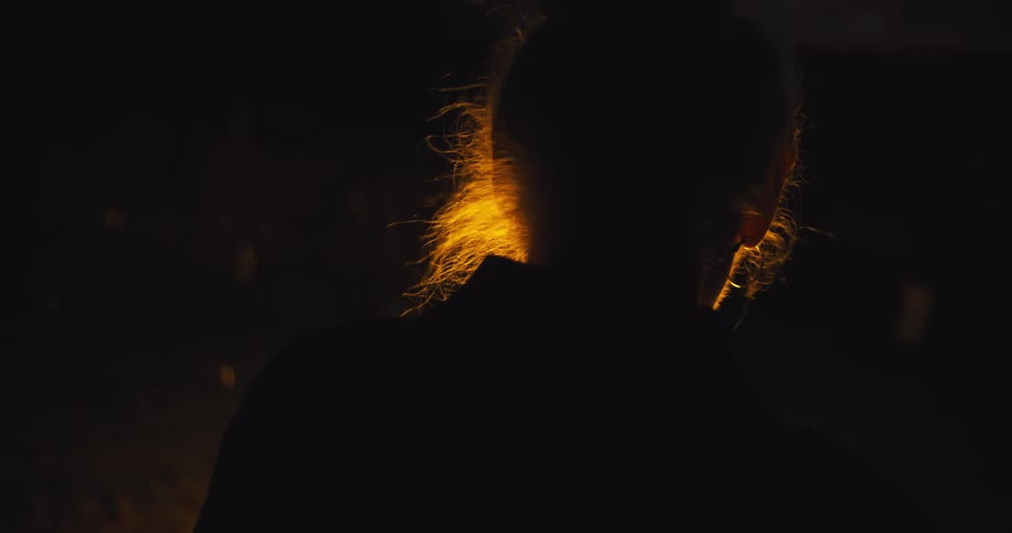A very close view shows the silhouette of a person from behind with their long light colored hair illuminated with a warm orange glow of a campfire