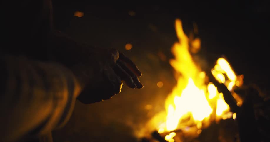Based on the video a closeup shot shows the hands of a person warming themselves by a bonfire at night the flames are bright and cast a warm glow on their hands and the surroundings