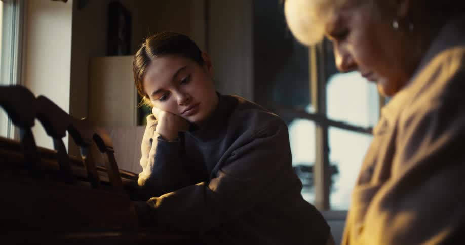A teenage girl with her hand on her chin watches an older woman play the piano