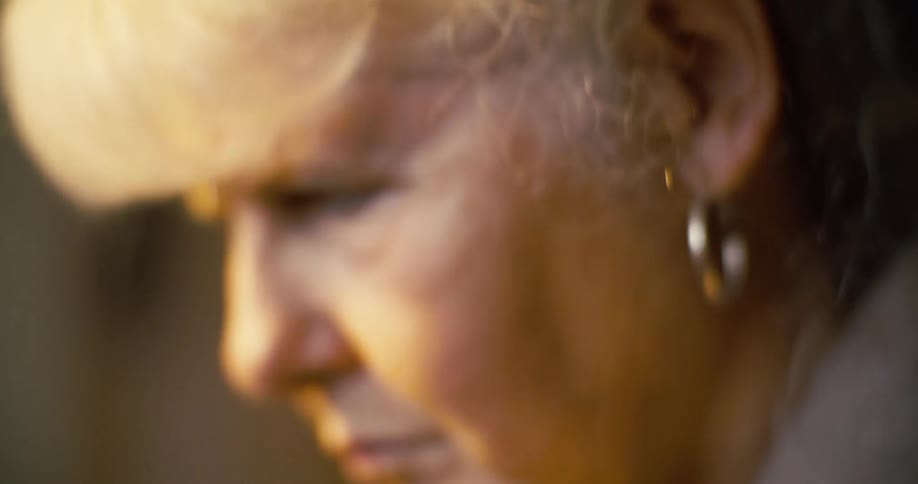 A close view of a woman with white hair and a large hoop earring shows her looking down with a serious expression with light from the left side of her face revealing the wrinkles around her eye and mouth