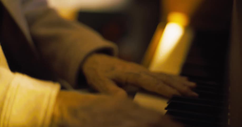 The video shows a closeup of a persons hands with wrinkled skin playing the keys of a piano the lighting is warm and dim creating a soft blurry background