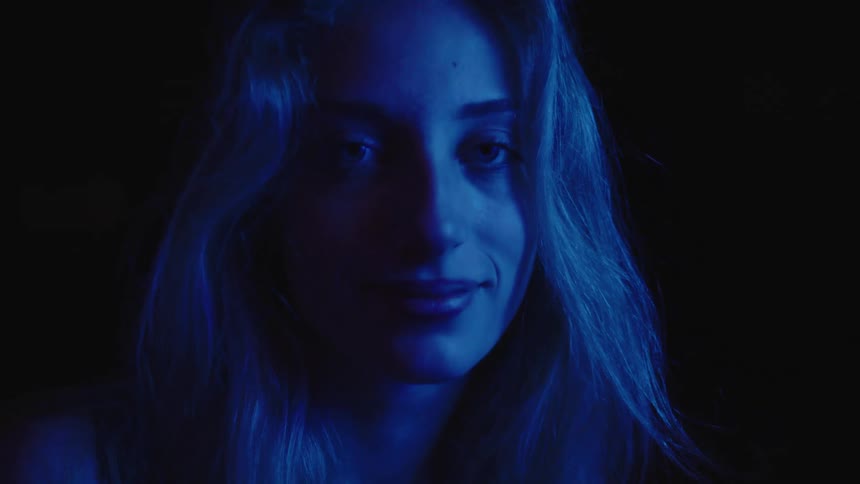 A close view shows a woman with long light colored hair with her head down and a slight smile as a blue light illuminates the right side of her face