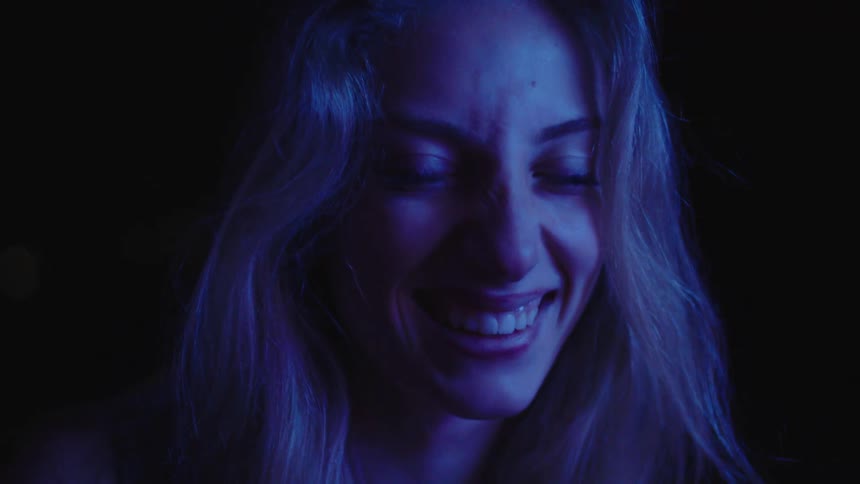 A woman with light colored hair and a bright confident smile is seen in a close view lit by a cool blue light from the right side of her face as she looks down and then back toward the camera