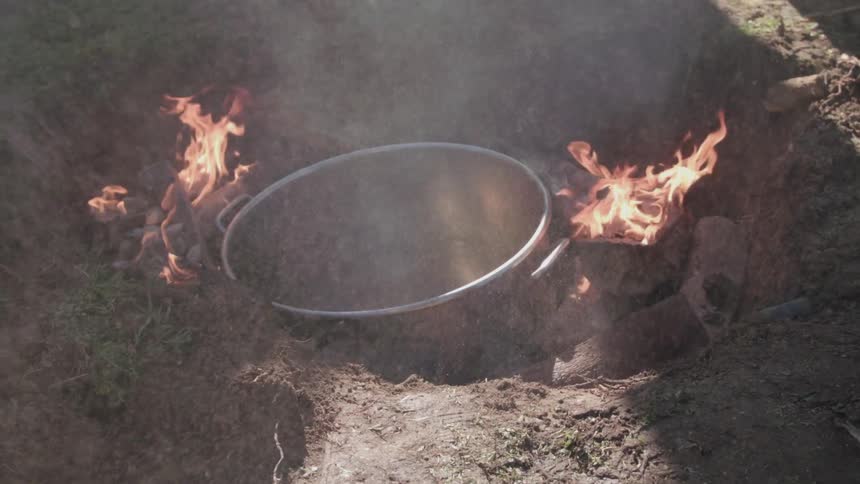 A large pot cooks on an outdoor fire as a person watches