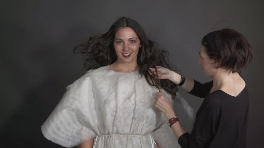 A woman ratting the hair of another woman who has long, brunette hair and is wearing a white dress as her hair is blowing back in a breeze