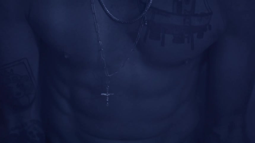 A close view shows a man with several tattoos on his torso and arms wearing multiple silver chains with a cross pendant