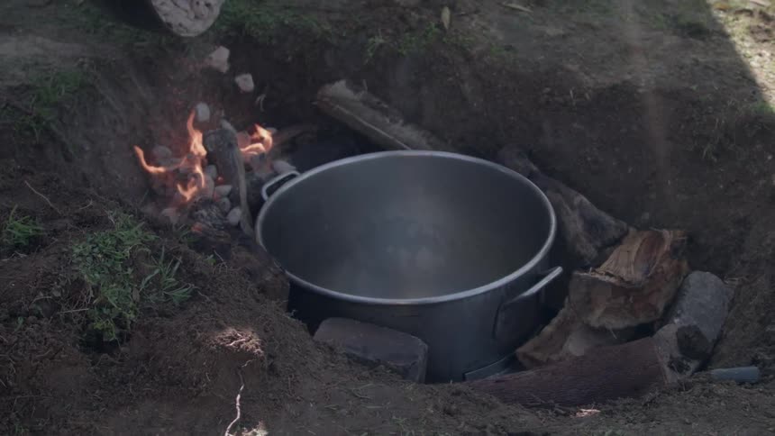 A large pot is in a hole in the ground and someone is pouring hot coals down around it