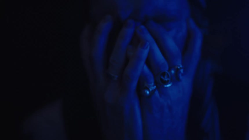 A young man wearing several rings and a watch sits in a dark room looking sad illuminated by dim blue light