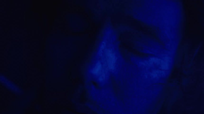 A close view shows a person with a beard and mustache with a very bright blue light shining across the right side of his face as he blows smoke to the camera
