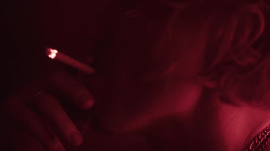 A close view of a woman smoking a lit cigarette is shown with her neck and mouth visible from the side and a necklace around her neck all lit by a red light