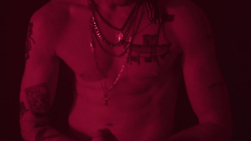 The video shows a man with a bare torso and multiple tattoos on his chest arms and stomach he is wearing several necklaces and bracelets and is bathed in a deep red light