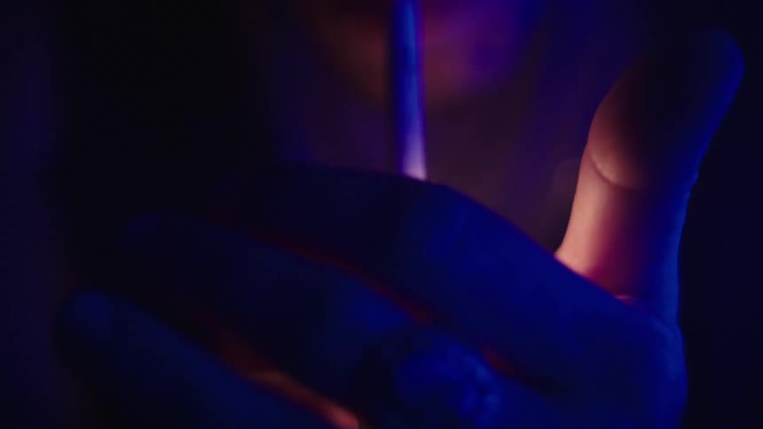 A man in a dark room illuminated by blue fluorescent light smokes a cigarette