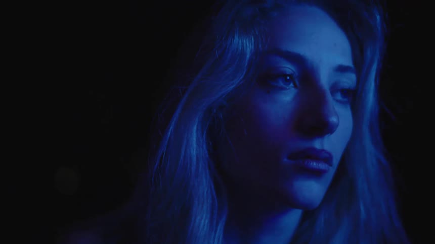 The video shows a woman with long blonde hair and a somber expression illuminated by a pulsing blue light she is seen looking down and then slowly turning her head to the side