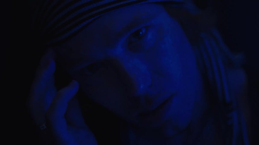 A man looks directly at the camera in dark blue light