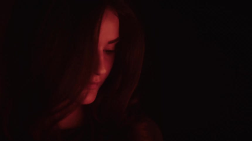 A close view of a woman with long dark hair with her head down and her eyes looking up to her left with the left side of her face illuminated by a deep orange light while the rest of the scene is dark